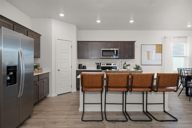 kitchen with light hardwood / wood-style floors, stainless steel appliances, dark brown cabinetry, a breakfast bar, and a kitchen island with sink