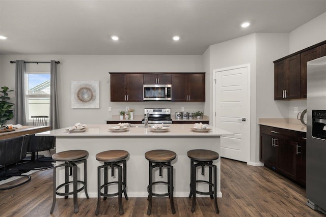 kitchen with dark hardwood / wood-style flooring, a kitchen bar, a kitchen island with sink, dark brown cabinets, and appliances with stainless steel finishes