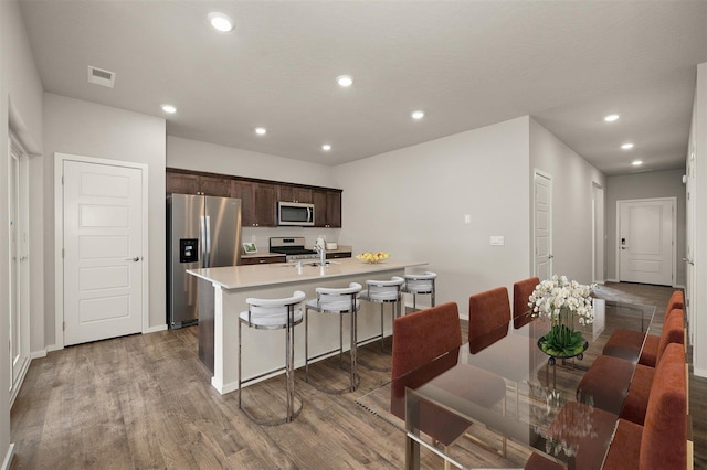 kitchen featuring a center island with sink, a kitchen breakfast bar, dark brown cabinetry, wood-type flooring, and stainless steel appliances