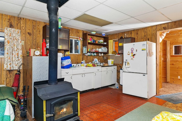 kitchen featuring a drop ceiling, wood walls, a wood stove, white refrigerator, and white cabinetry