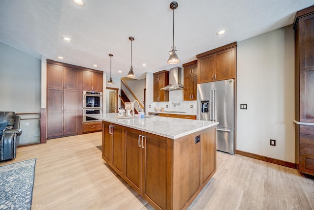 kitchen featuring wall chimney exhaust hood, stainless steel appliances, light stone counters, pendant lighting, and a center island with sink