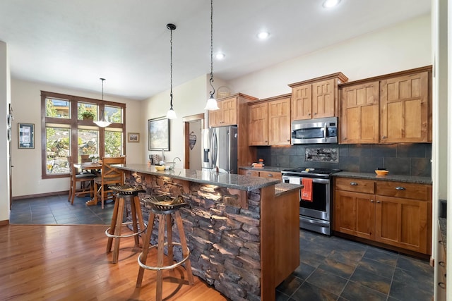 kitchen featuring appliances with stainless steel finishes, backsplash, a kitchen breakfast bar, a kitchen island with sink, and decorative light fixtures