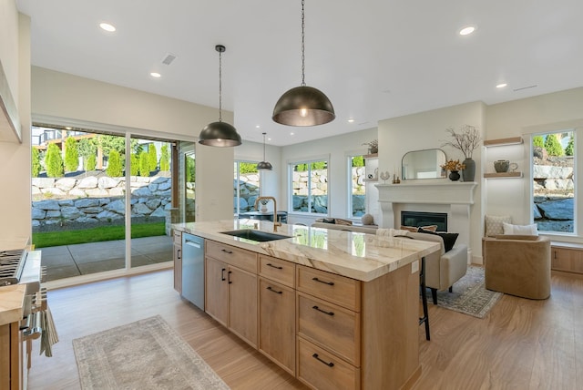 kitchen featuring light brown cabinets, a center island with sink, dishwasher, a glass covered fireplace, and a sink