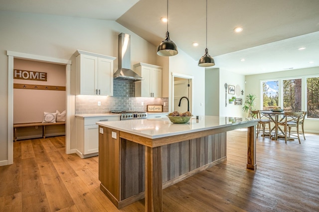 kitchen featuring stainless steel stove, vaulted ceiling, wall chimney exhaust hood, an island with sink, and white cabinetry