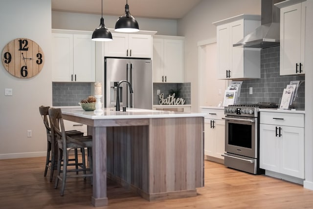 kitchen with wall chimney range hood, pendant lighting, a center island with sink, white cabinets, and high end appliances