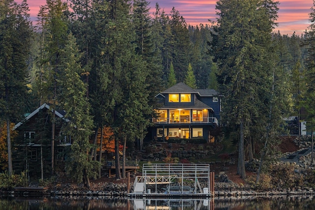 back of house at dusk featuring a balcony, a forest view, and a water view