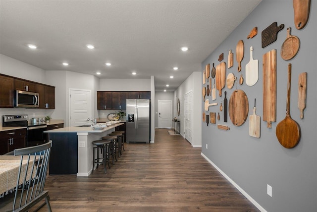 kitchen with stainless steel appliances, a kitchen breakfast bar, dark brown cabinets, dark hardwood / wood-style floors, and a kitchen island with sink