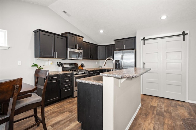 kitchen featuring lofted ceiling, tasteful backsplash, an island with sink, a barn door, and appliances with stainless steel finishes