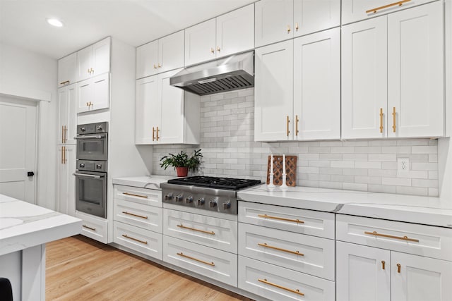 kitchen with white cabinets, backsplash, light stone countertops, exhaust hood, and appliances with stainless steel finishes