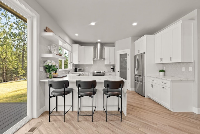 kitchen with a breakfast bar area, white cabinetry, stainless steel appliances, and wall chimney range hood