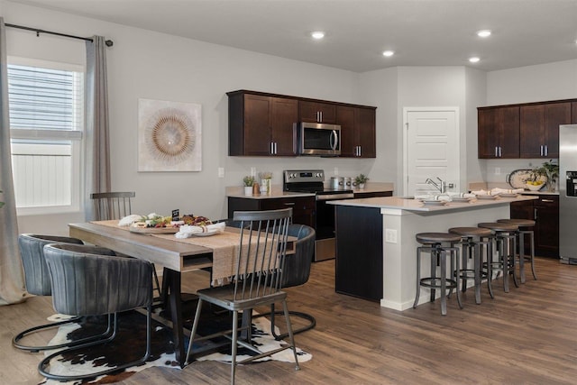 kitchen featuring dark brown cabinetry, dark wood-type flooring, stainless steel appliances, a center island with sink, and a breakfast bar area