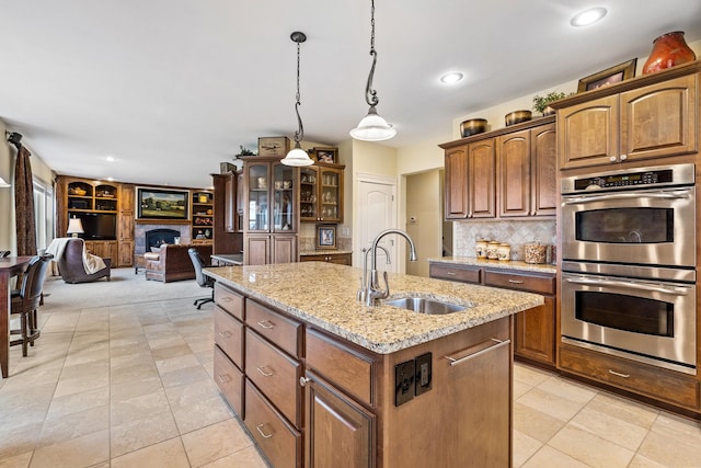 kitchen featuring decorative light fixtures, sink, an island with sink, stainless steel double oven, and light stone counters