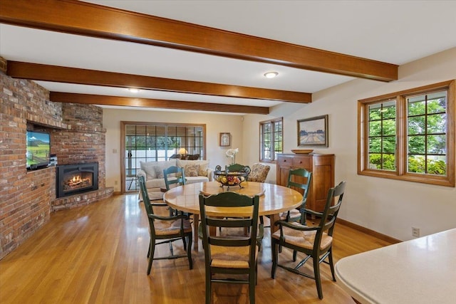 dining space featuring beamed ceiling, a healthy amount of sunlight, a brick fireplace, and light wood-type flooring