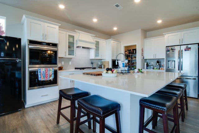 kitchen with stainless steel appliances, white cabinetry, custom range hood, and an island with sink