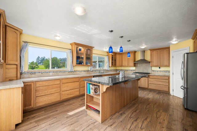 kitchen featuring wall chimney exhaust hood, wood-type flooring, a center island with sink, appliances with stainless steel finishes, and pendant lighting