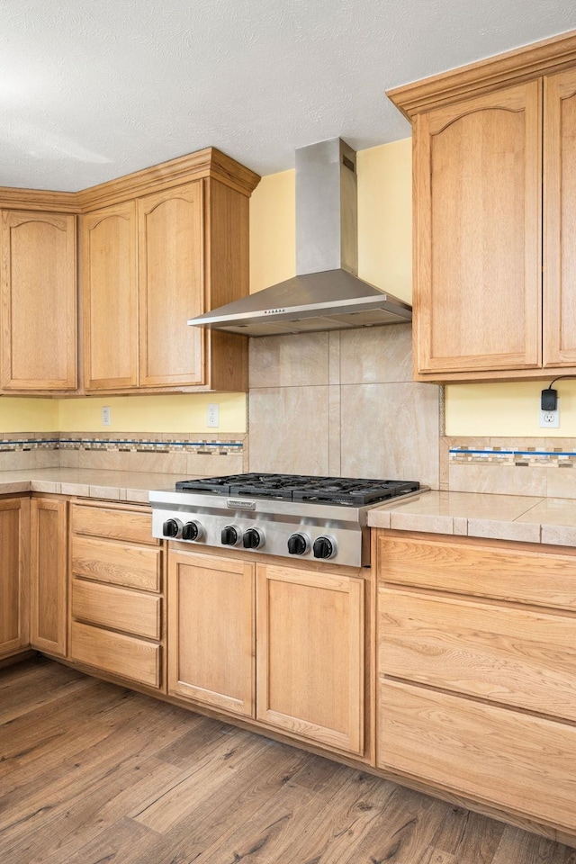 kitchen featuring wall chimney range hood, backsplash, tile counters, stainless steel gas stovetop, and light wood-type flooring