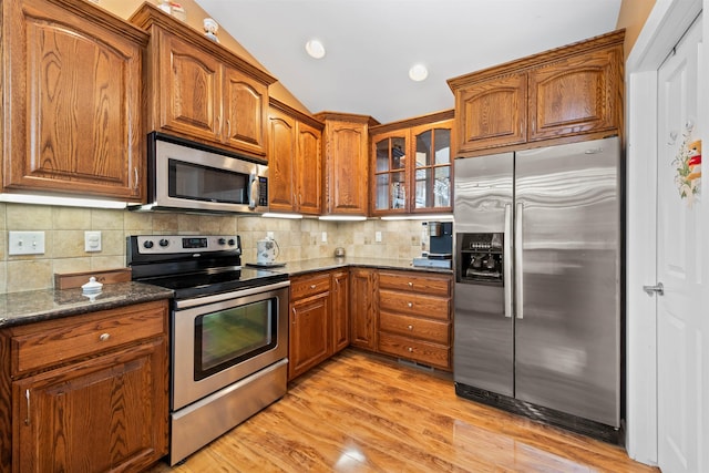 kitchen with appliances with stainless steel finishes, tasteful backsplash, lofted ceiling, dark stone countertops, and light wood-type flooring