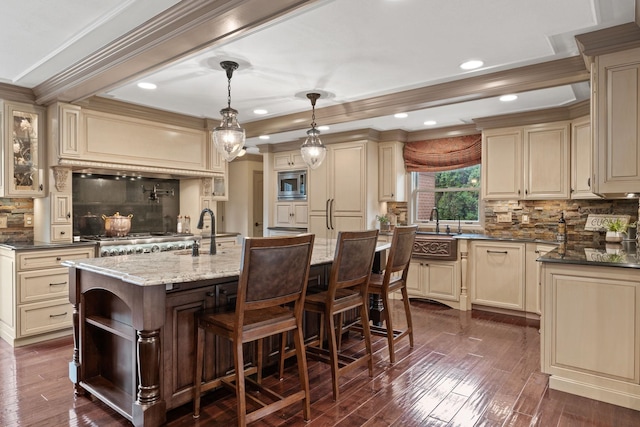 kitchen featuring stainless steel microwave, decorative light fixtures, dark stone counters, a center island with sink, and cream cabinetry