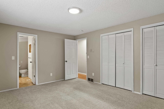 unfurnished bedroom featuring light colored carpet, ensuite bathroom, a textured ceiling, and two closets