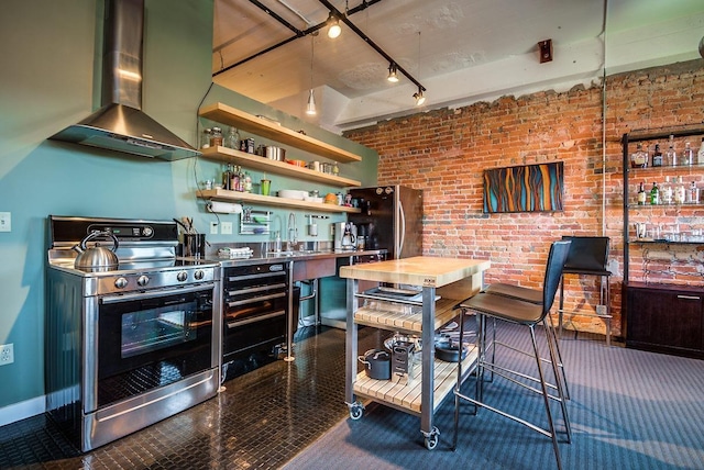 kitchen with brick wall, appliances with stainless steel finishes, rail lighting, and wall chimney range hood