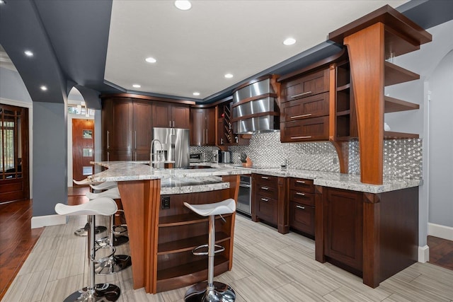 kitchen with wall chimney range hood, sink, a breakfast bar area, appliances with stainless steel finishes, and light stone counters