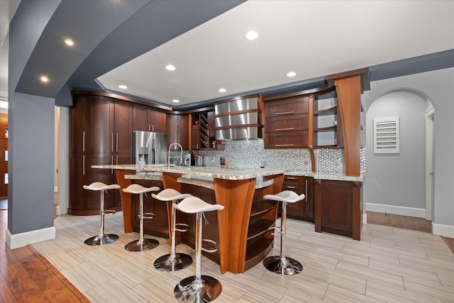 kitchen with a kitchen breakfast bar, stainless steel refrigerator with ice dispenser, light stone countertops, decorative backsplash, and wall chimney exhaust hood