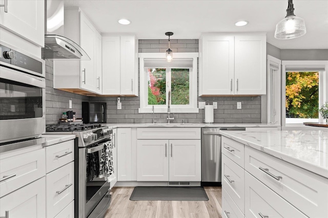 kitchen with wall chimney range hood, sink, white cabinetry, stainless steel appliances, and decorative light fixtures