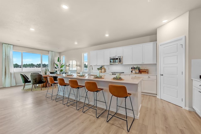 kitchen with light countertops, stainless steel microwave, a center island with sink, and white cabinets