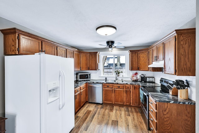 kitchen with under cabinet range hood, stainless steel appliances, light wood-type flooring, brown cabinetry, and dark stone countertops