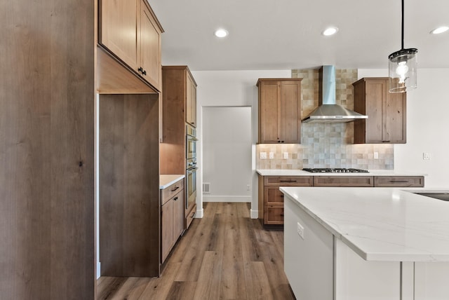 kitchen with pendant lighting, light countertops, appliances with stainless steel finishes, brown cabinetry, and wall chimney range hood