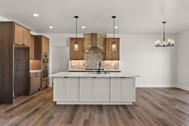 kitchen with a center island with sink, brown cabinetry, wall chimney exhaust hood, light countertops, and pendant lighting