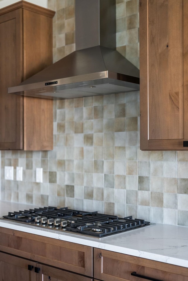 kitchen featuring wall chimney range hood, brown cabinetry, light stone counters, and decorative backsplash