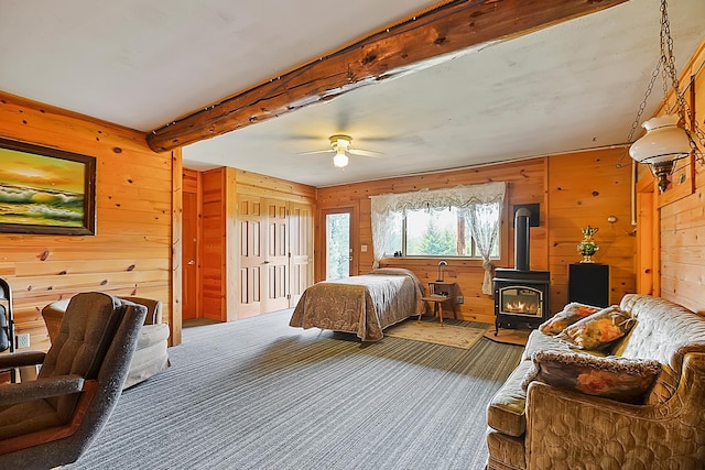 carpeted bedroom featuring a ceiling fan, a wood stove, beamed ceiling, and wooden walls