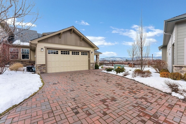 snow covered property featuring a garage, stone siding, decorative driveway, and board and batten siding