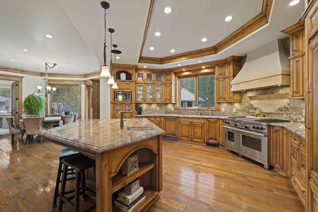 kitchen with a kitchen island with sink, premium range hood, appliances with stainless steel finishes, brown cabinets, and glass insert cabinets