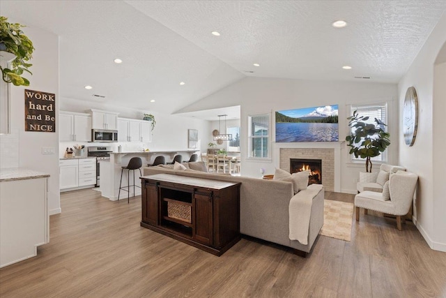 living room featuring light wood finished floors, a warm lit fireplace, lofted ceiling, a textured ceiling, and recessed lighting