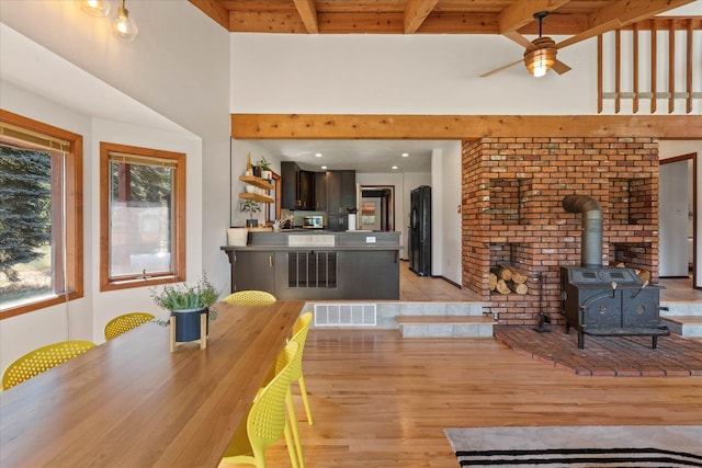 dining room featuring a high ceiling, visible vents, light wood-style floors, beamed ceiling, and a wood stove