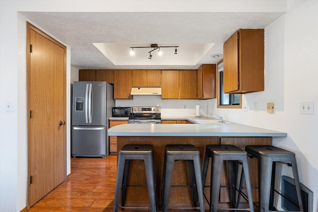 kitchen featuring stainless steel appliances, brown cabinetry, a textured ceiling, a peninsula, and under cabinet range hood