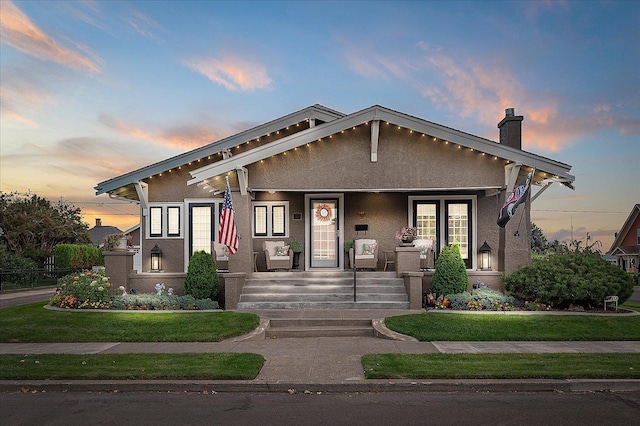 view of front facade with covered porch, a chimney, a lawn, and stucco siding