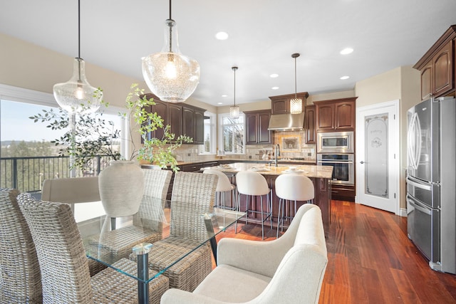 kitchen with appliances with stainless steel finishes, dark wood-style floors, wall chimney exhaust hood, tasteful backsplash, and a center island with sink