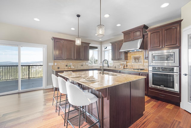 kitchen featuring light stone counters, appliances with stainless steel finishes, a kitchen island with sink, a sink, and under cabinet range hood