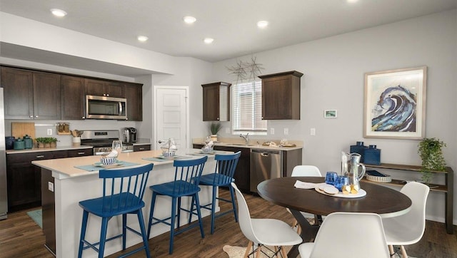 kitchen with stainless steel appliances, an island with sink, dark brown cabinetry, and dark wood-style flooring