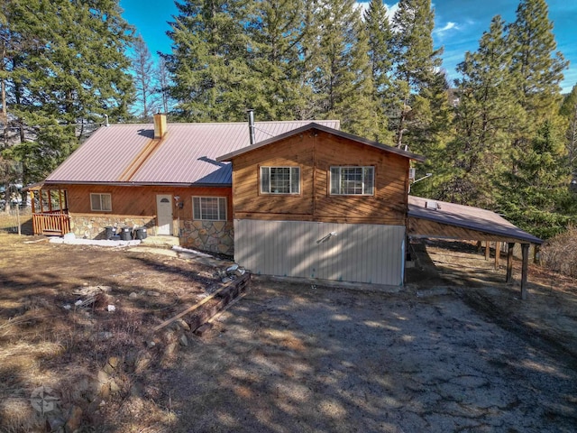 back of house with stone siding, a chimney, metal roof, and a carport