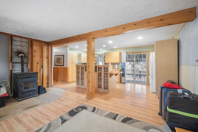 living room featuring light wood finished floors, wooden walls, a wood stove, a textured ceiling, and beam ceiling