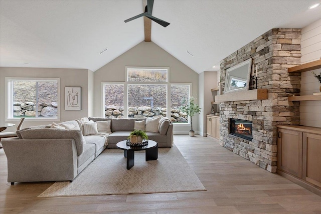 living area with light wood-type flooring, ceiling fan, a stone fireplace, and lofted ceiling with beams