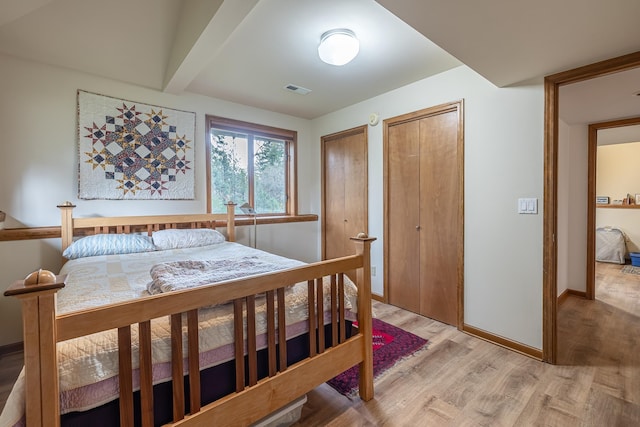 bedroom with baseboards, visible vents, light wood-style flooring, beamed ceiling, and two closets