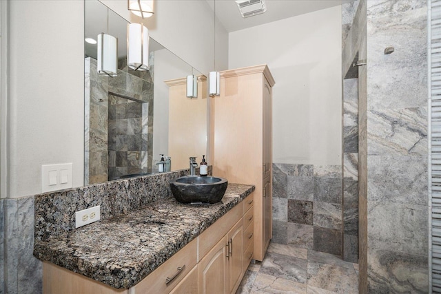 bathroom with visible vents, stone finish floor, a tile shower, vanity, and tile walls