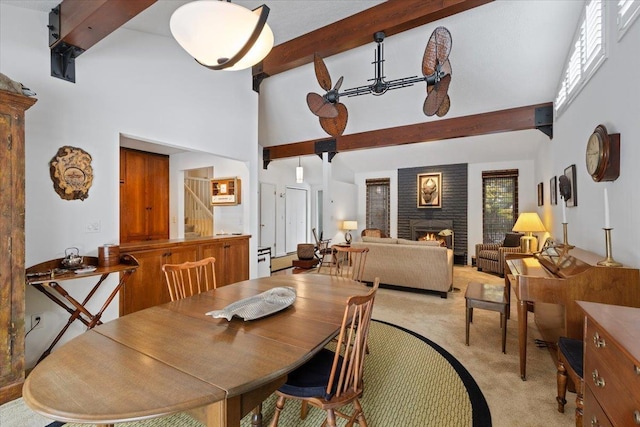 dining space featuring light carpet, a warm lit fireplace, high vaulted ceiling, and beam ceiling