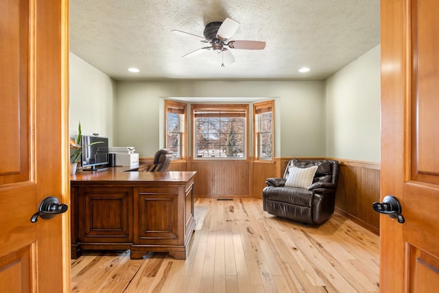 office area with a wainscoted wall, light wood finished floors, a ceiling fan, wooden walls, and a textured ceiling