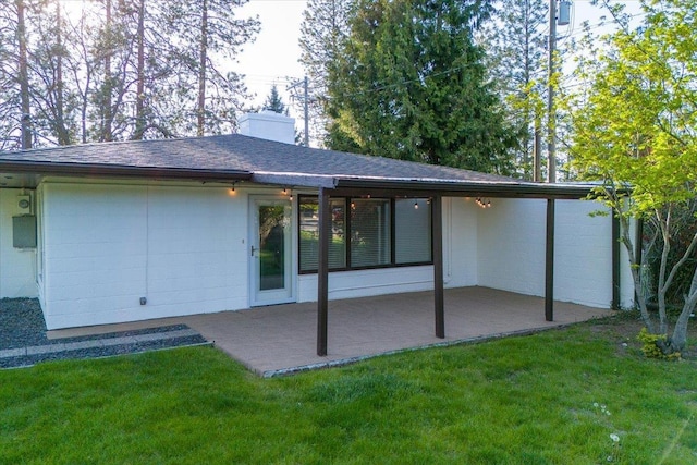 back of property with a yard, a shingled roof, a chimney, and a patio